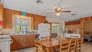 A compact kitchen with wooden cabinets, a dining table with chairs, a ceiling fan, and a view through the window.