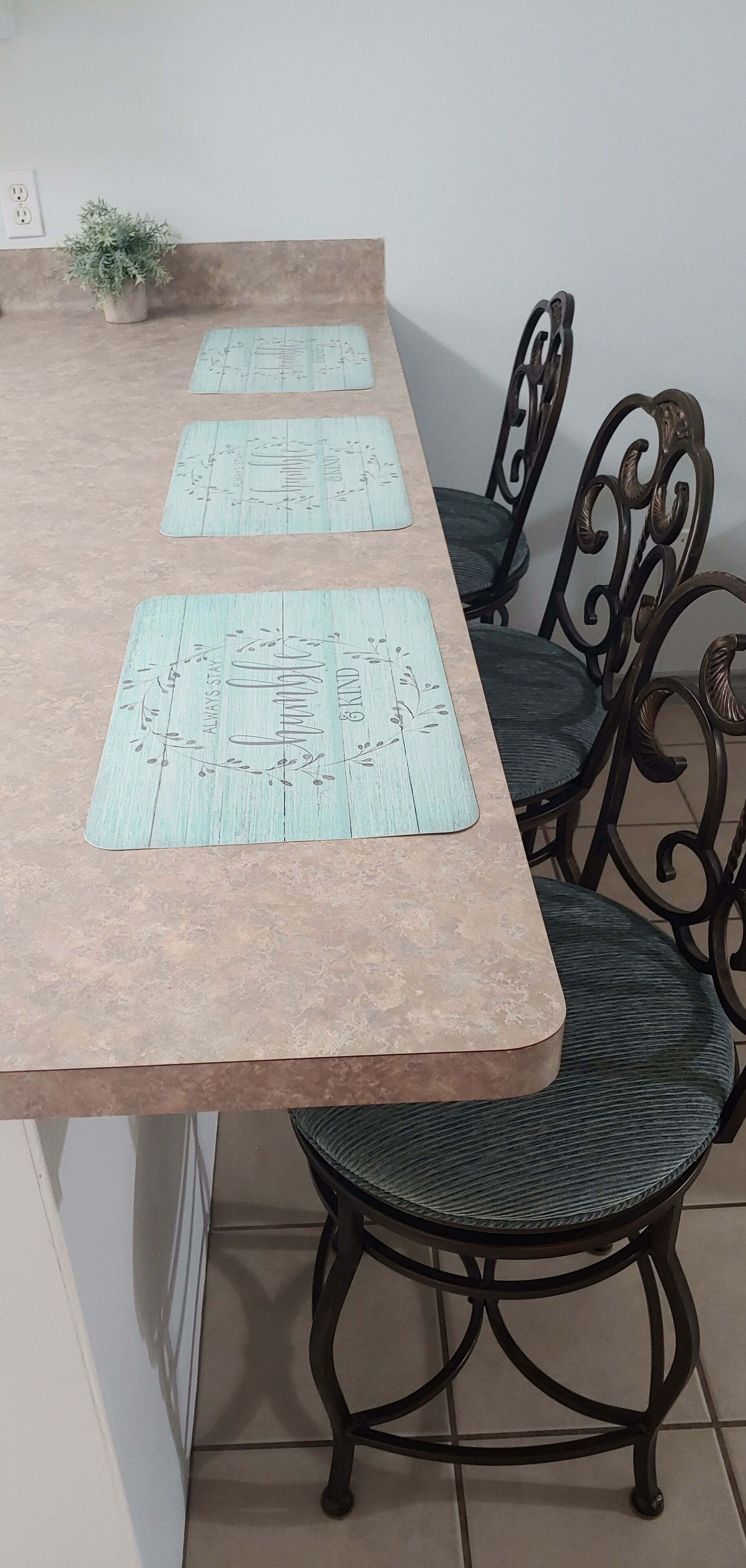 Mesa de jantar com superfície marrom-clara e três jogos americanos com motivos florais.