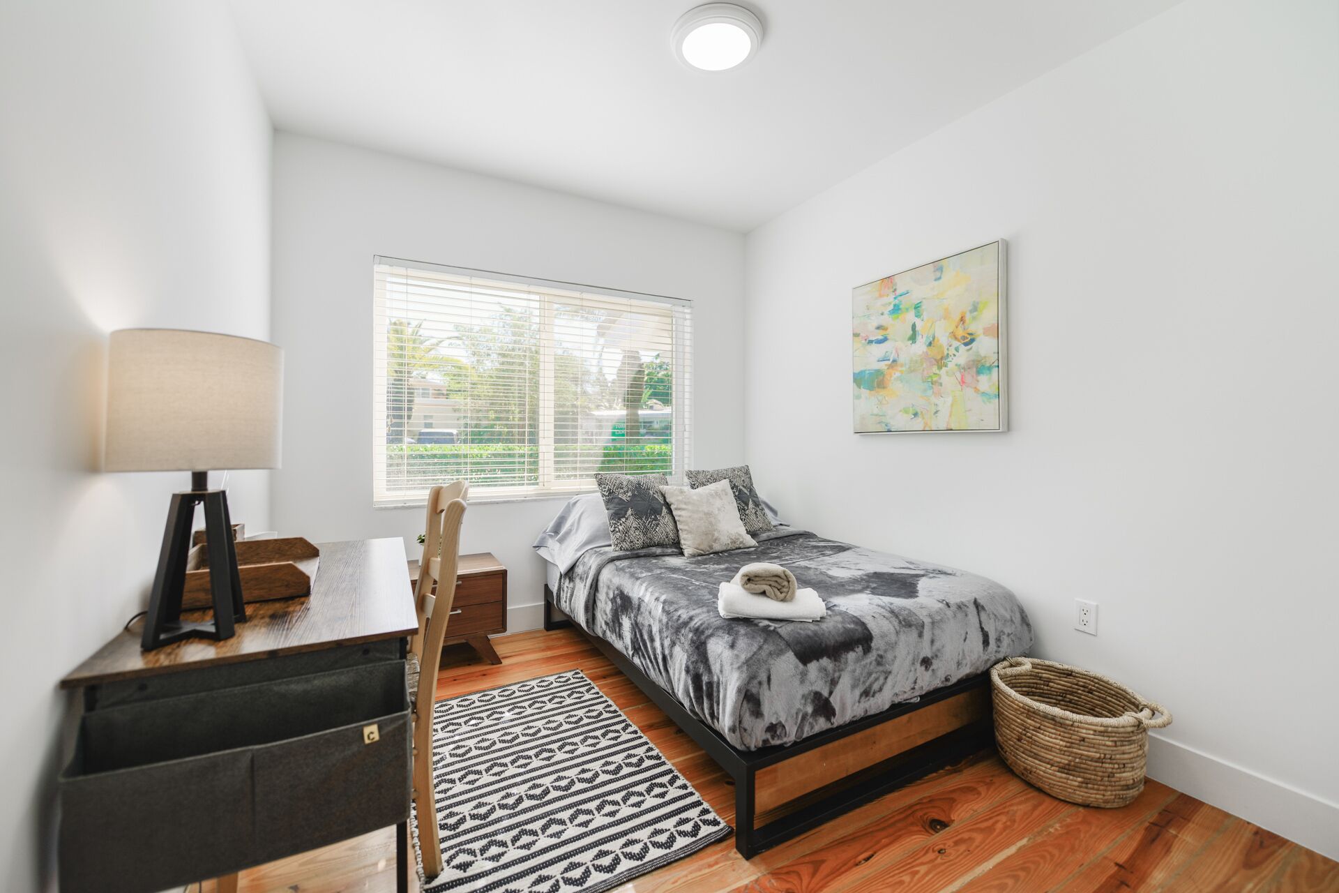 A bedroom with a bed, a desk, a chair, a lamp, a window, and a painting.