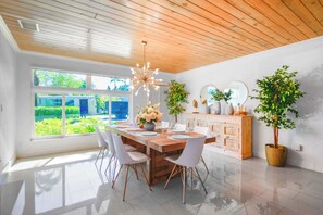 Un comedor con techo de madera, un ventanal con vistas a follaje, una lámpara moderna y una mesa de comedor blanca con sillas.