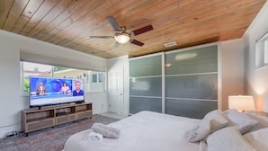 Ein Schlafzimmer mit einem großen Bett, einem Fernseher, der eine Nachrichtensendung zeigt, einem Deckenventilator und einer Schiebetür zum Balkon.
