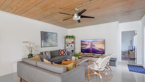 Ein modernes Wohnzimmer mit einem grauen Sofa, einem Holztisch, einem Fernseher der eine Wüstenstraße zeigt, einem Deckenventilator und einem Bücherregal mit Pflanzen.
