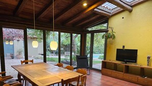 A spacious dining area with a large wooden table and chairs, a television on a wooden stand, and a view of the garden through large glass doors. - Casa Las Colinas by Vilvey Turismo (Chacras de Coria)
