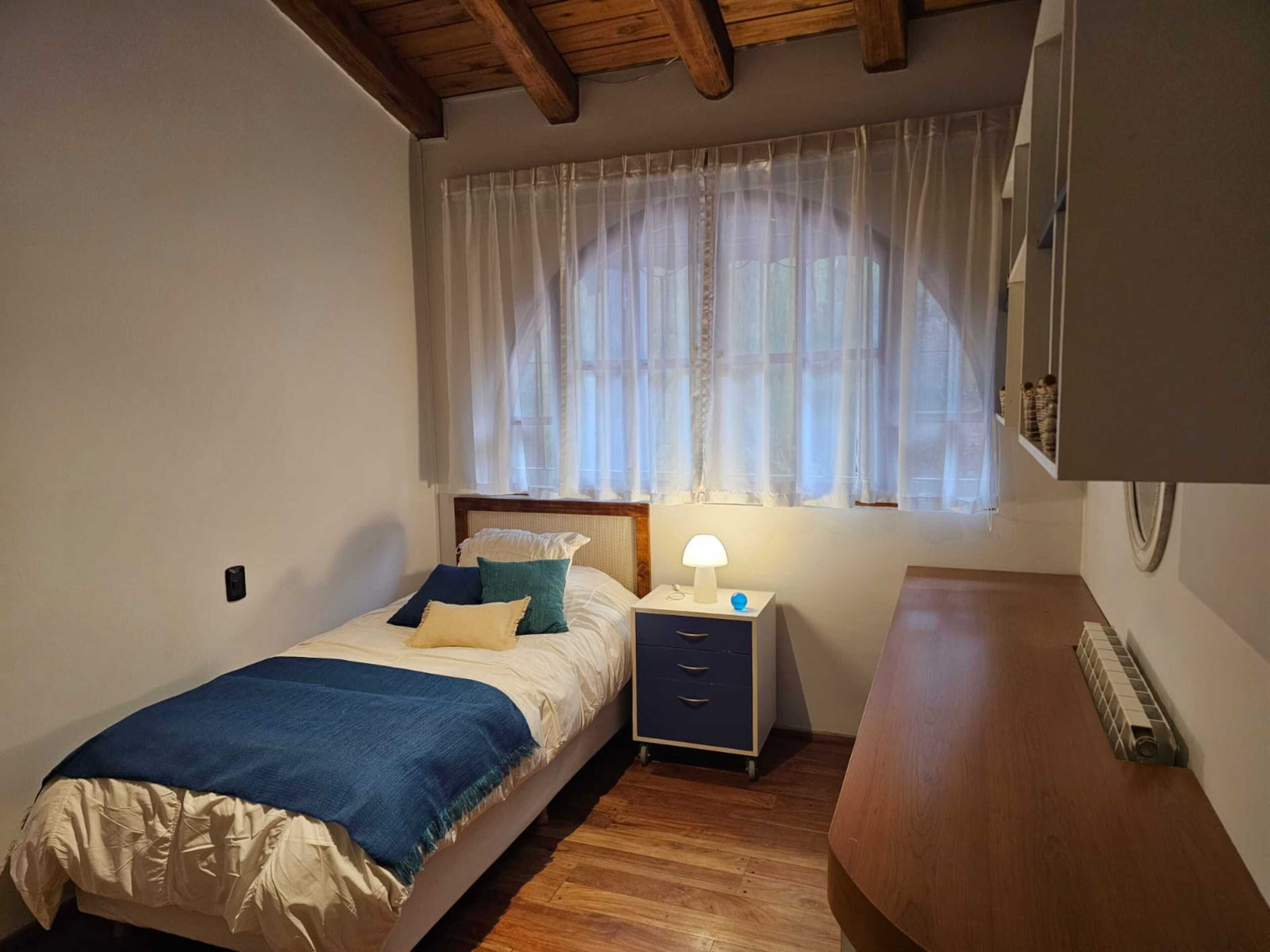 A bedroom with a bed, bedside table, lamp, and a window with sheer curtains.