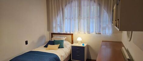 A bedroom with a bed, bedside table, lamp, and a window with sheer curtains.
