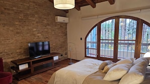 A bedroom with a brick wall, wooden floor, a TV on a wooden stand, a bed with pillows, a red chair, and a large arched window. - Casa Las Colinas by Vilvey Turismo (Chacras de Coria)