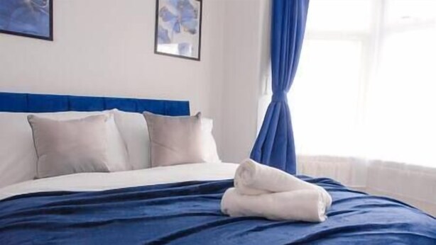 Ein ordentlich bezogenes Bett mit blauer Bettdecke und weißen Kissen, ein blauer Kopfteil und ein Fenster mit blauen Vorhängen.