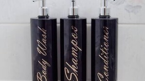 Drei schwarze Flaschen mit Dosierern, beschriftet mit 'Body Wash', 'Shampoo' und 'Conditioner', auf einer weißen Oberfläche.