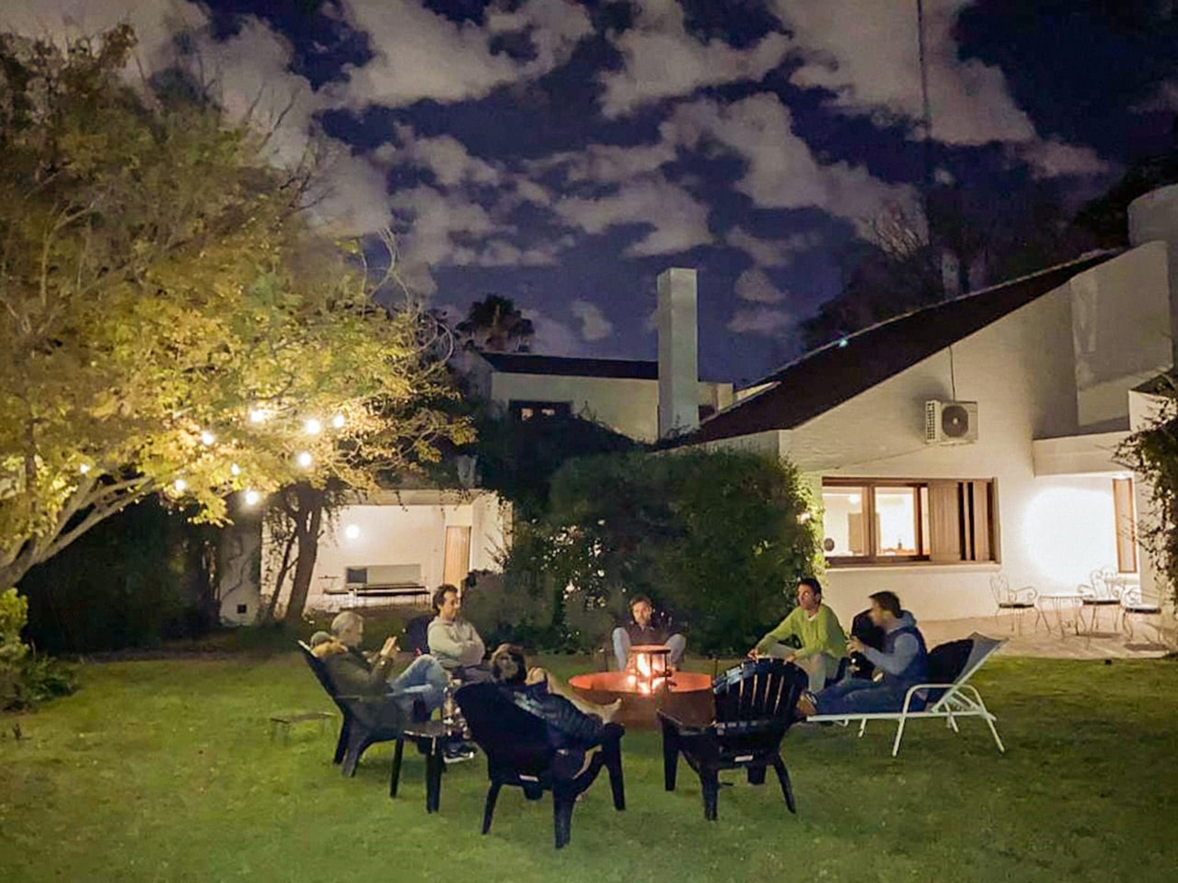 A group of people gathered around a fire pit in a backyard with a modern house in the background.