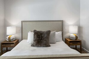 A neatly made bed with a grey fluffy pillow, two bedside tables with lamps, and a plain wall.