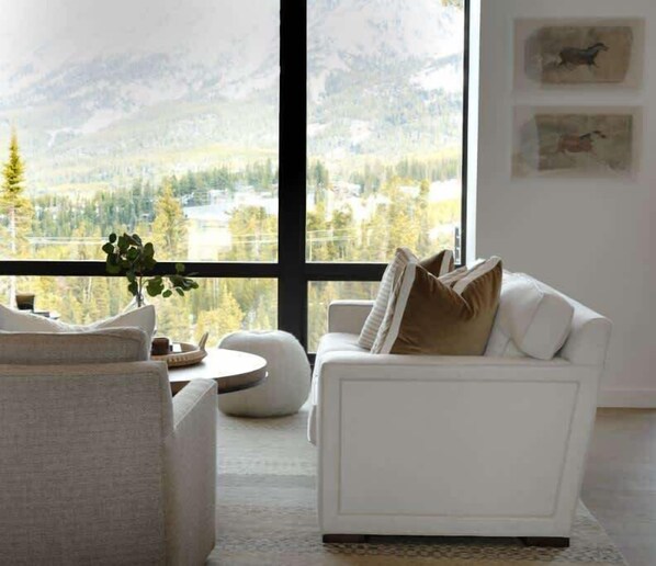 A modern living room with a white sofa, a round coffee table, and a view of mountains through large windows.