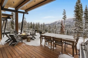 A wooden deck with outdoor furniture, a fire pit, and a view of snow-covered trees and mountains. - Luxury Ski-In/Ski-Out Moonlight Basin Home (Big Sky)