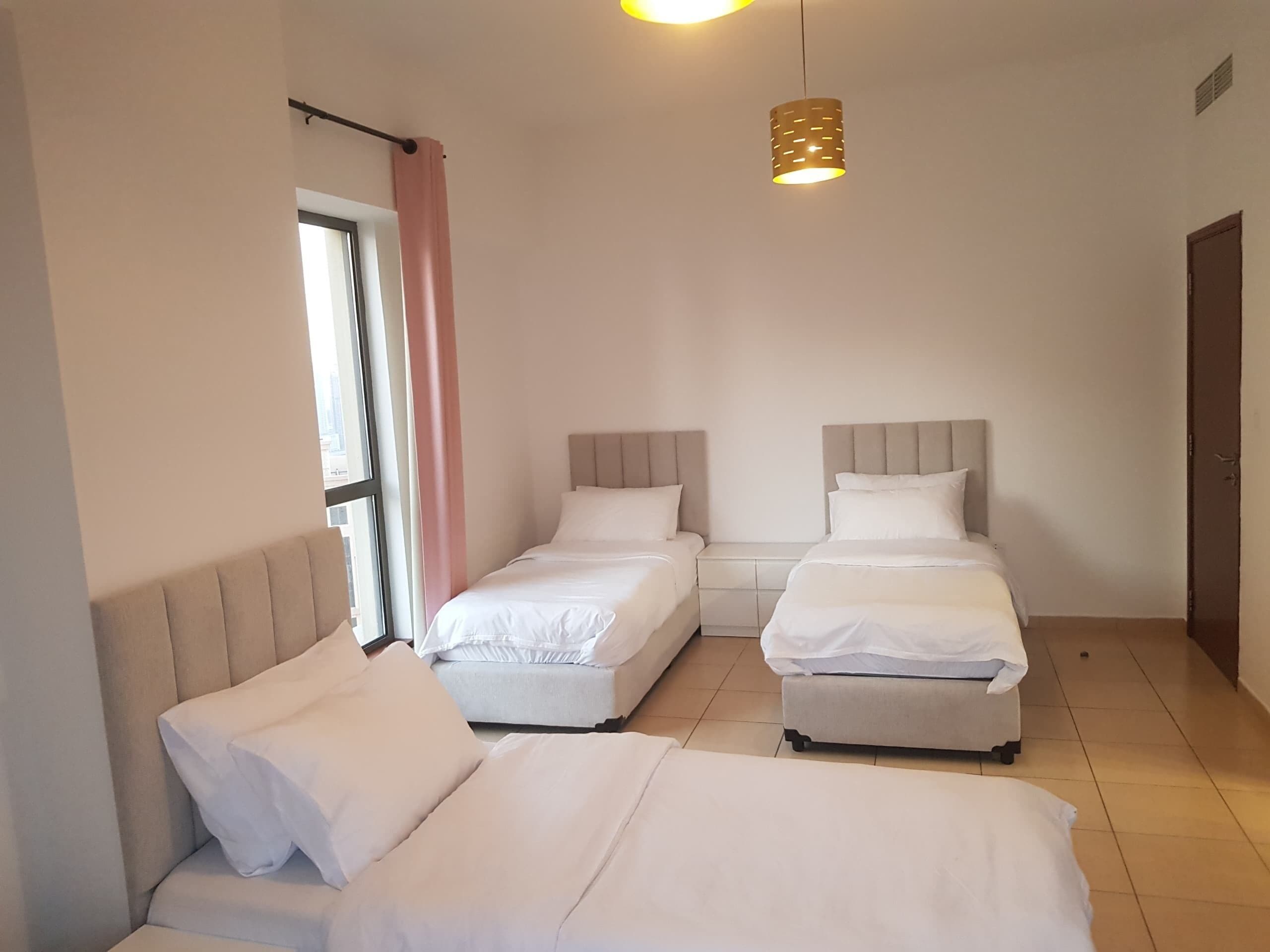 A hotel room with two single beds, a sofa, a window with curtains, and a hanging light fixture.