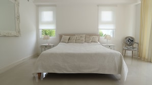 A large bed with a white quilt, two bedside tables with lamps, a chair, and a window with blinds. - Las Brísas Villa - Chateau Elysium (Mahé Island)