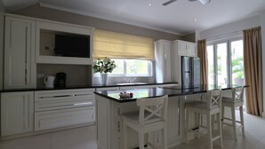 A modern kitchen with white cabinetry, a black countertop, a built-in oven, and a refrigerator. There is a window with a view of greenery outside. - Las Brísas Villa - Chateau Elysium (Mahé Island)
