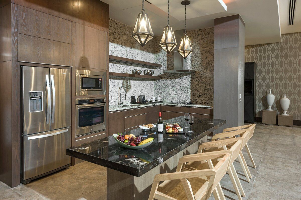 A modern kitchen with stainless steel appliances, a marble island, wooden chairs, and pendant lighting.