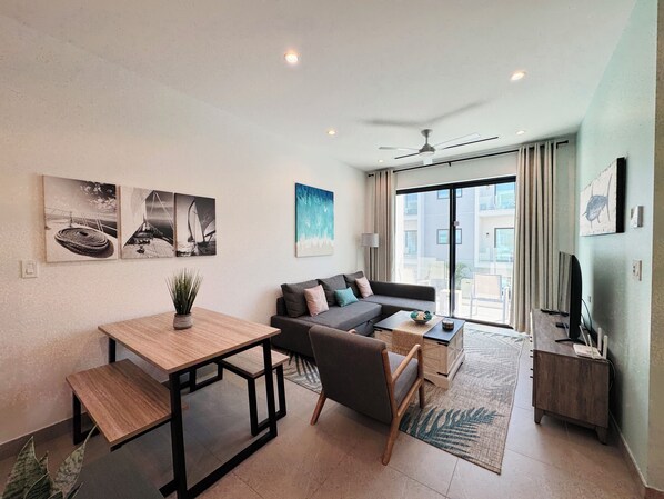 A modern living room with a sofa, coffee table, and a dining table with chairs. There are artworks on the walls, a ceiling fan, and a view of the outside balcony.
