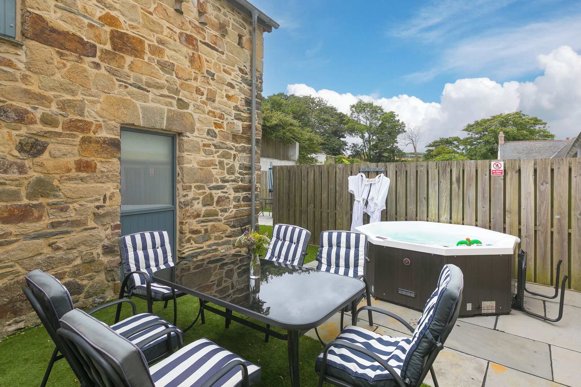 An outdoor patio with a stone wall, a hot tub, striped lounge chairs, and a wooden fence.