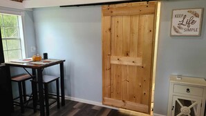 A room with a wooden door, a small table with a chair, a window, and a wall with a 'KEEP Life Simple' sign. - Newly transformed farm cottage off the beaten path! (Minford)