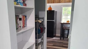 A narrow hallway with a white shelving unit on the left, a black refrigerator in the center, and a wooden table with chairs in the background. - Newly transformed farm cottage off the beaten path! (Minford)