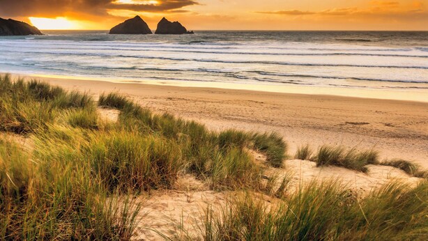 Una playa con dunas de arena y hierba en primer plano, y un atardecer sobre el océano con dos formaciones rocosas a lo lejos.