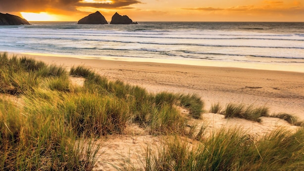 A beach with sand dunes and grass in the foreground, and a sunset over the ocean with two rock formations in the distance. - Carines Barns - Tre (Newquay)