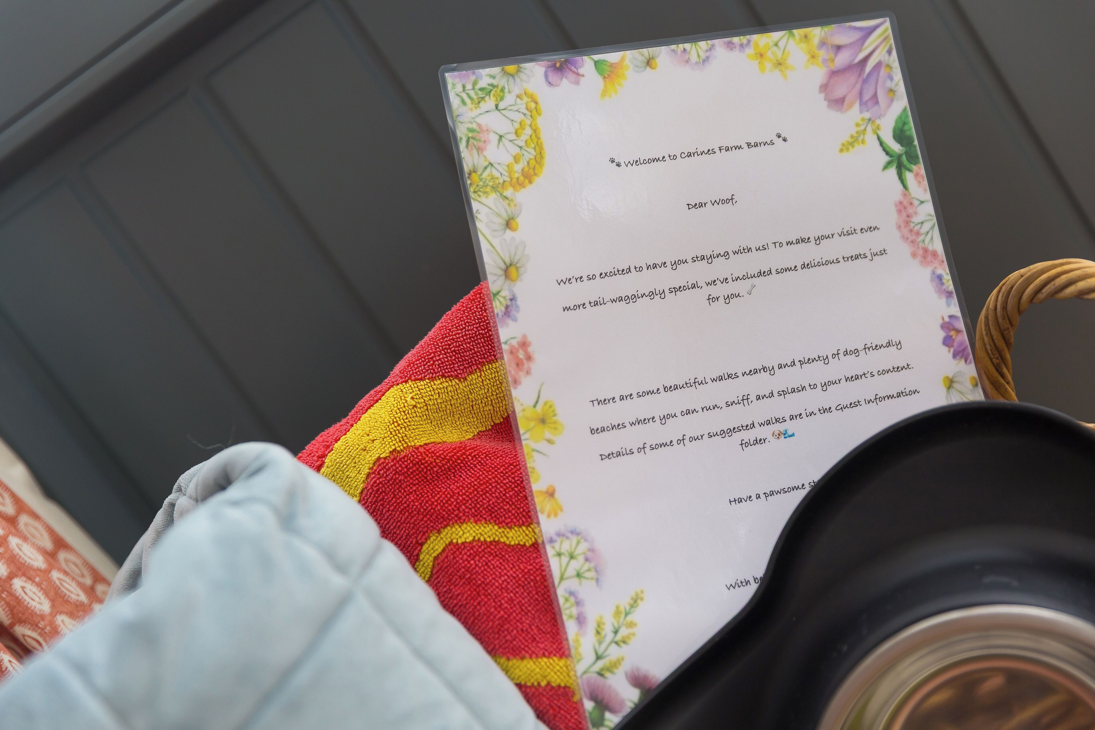 Tarjeta de bienvenida con diseño floral y texto, sobre una mesa junto a un cojín rojo y amarillo con estampado y un objeto negro.