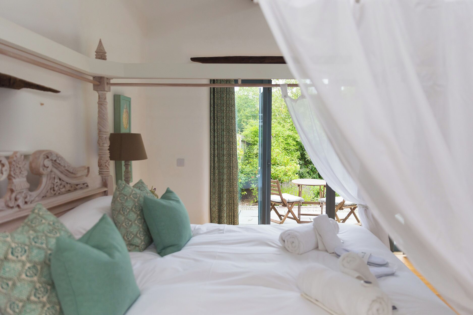 A bedroom with a bed, pillows, a lamp, and a view of an outdoor area with a table and chairs.