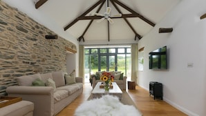 A spacious living room with a stone wall, wooden beams, a large window, a sofa, a coffee table, a fluffy white rug, and a fireplace. - Carines Barns - Wesley Barn (Newquay)