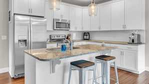 Une cuisine moderne avec des meubles blancs, des appareils en acier inoxydable, un plan de travail en granit et deux tabourets de bar.