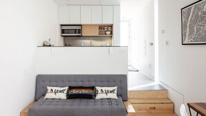 A modern living room with a grey sofa, a woven pouf, and a wooden staircase leading to a kitchen area. - Kasita Nest is a prefabricated, smart home for romantic getaways. (Austin)