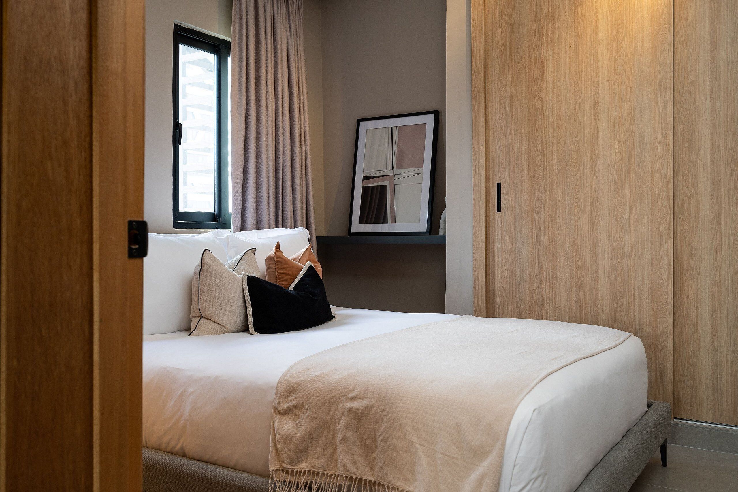 Una cama bien hecha con una manta color beige y almohadas, una ventana con cortinas y un cuadro en la pared.