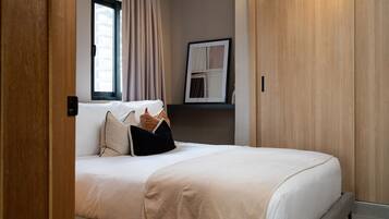 Una cama bien hecha con una manta color beige y almohadas, una ventana con cortinas y un cuadro en la pared.