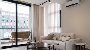 Un salon moderne avec un canapé, une table basse en verre, un fauteuil en osier et une vue sur un immeuble à travers de grandes fenêtres.