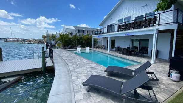 Un área de piscina con sillones, un muelle y un edificio con balcón con vista al agua.