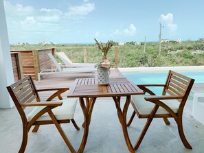 A wooden deck with a table and chairs overlooking a pool and a coastal view.
