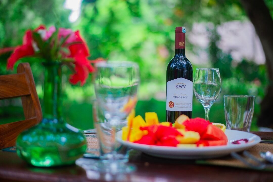 Eine Flasche KWV Pinotage Wein, ein Obstteller und zwei Gläser auf einem Tisch. Im Hintergrund ein Holzbstuhl und eine grüne Vase.