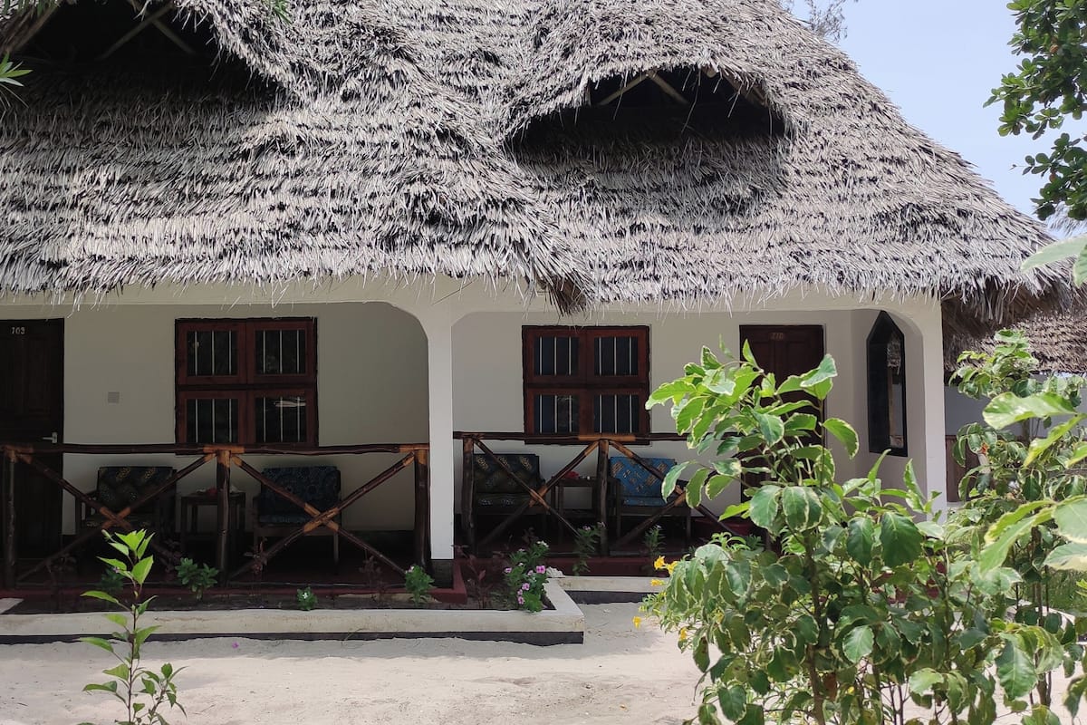 A traditional thatched-roof house with a porch, wooden furniture, and a number '703' on the door.