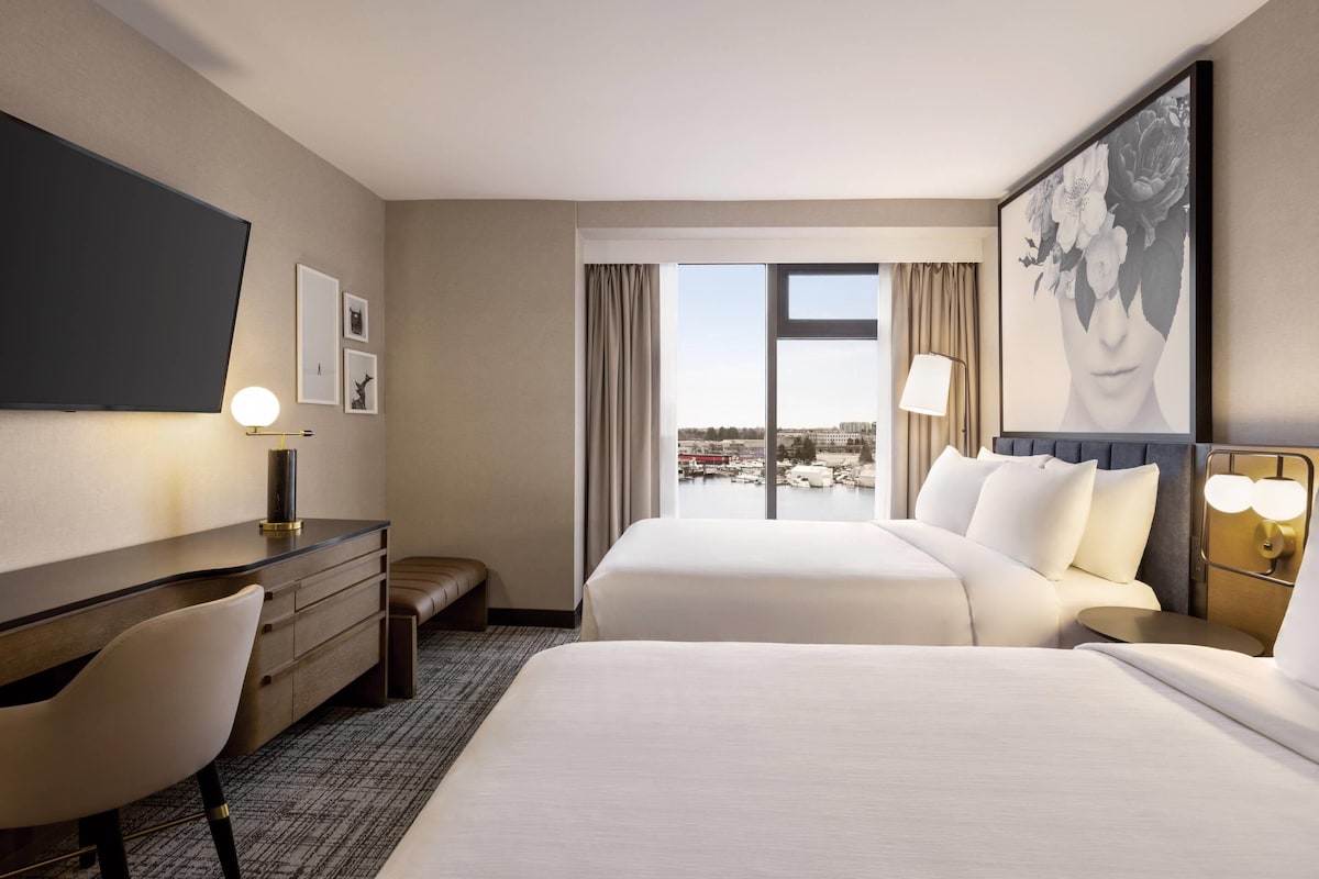 A hotel room with two beds, a desk, a chair, a TV, and a large window with a view of a marina.