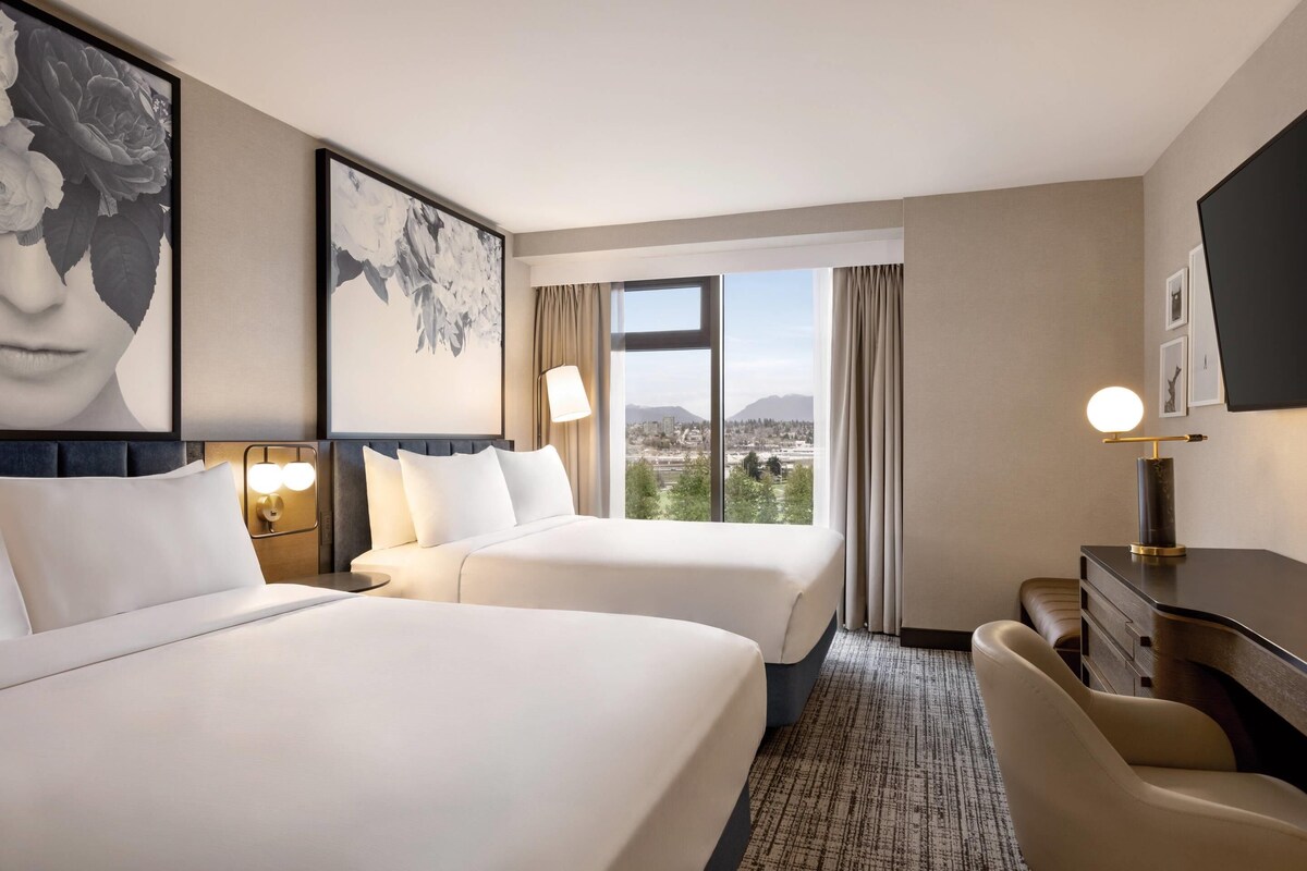 A hotel room with two beds, a desk, a chair, and a large window offering a city view.