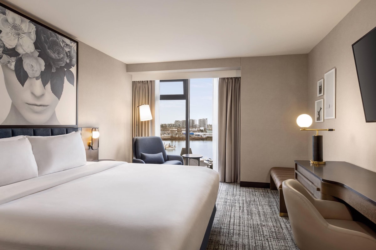 A hotel room with a large bed, a desk, a chair, a lamp, a picture on the wall, and a view of the city through the window.