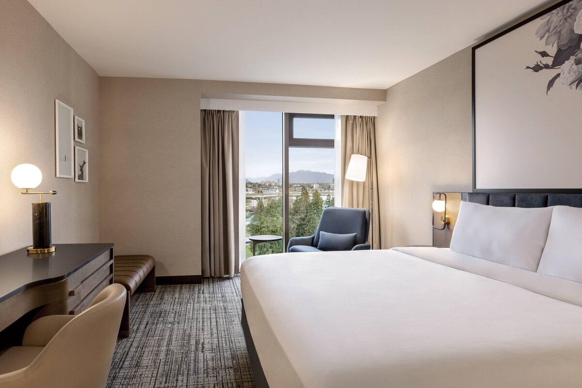 A hotel room with a large bed, a desk, a chair, and a view of the city through the window.