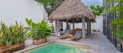 Un área de piscina con una estructura de techo de paja, sillas de descanso y plantas en macetas.