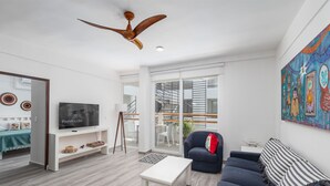 Ein modernes Wohnzimmer mit Flachbildfernseher, Deckenventilator, Sofa, Couchtisch und einem großen Fenster mit Blick ins Freie.