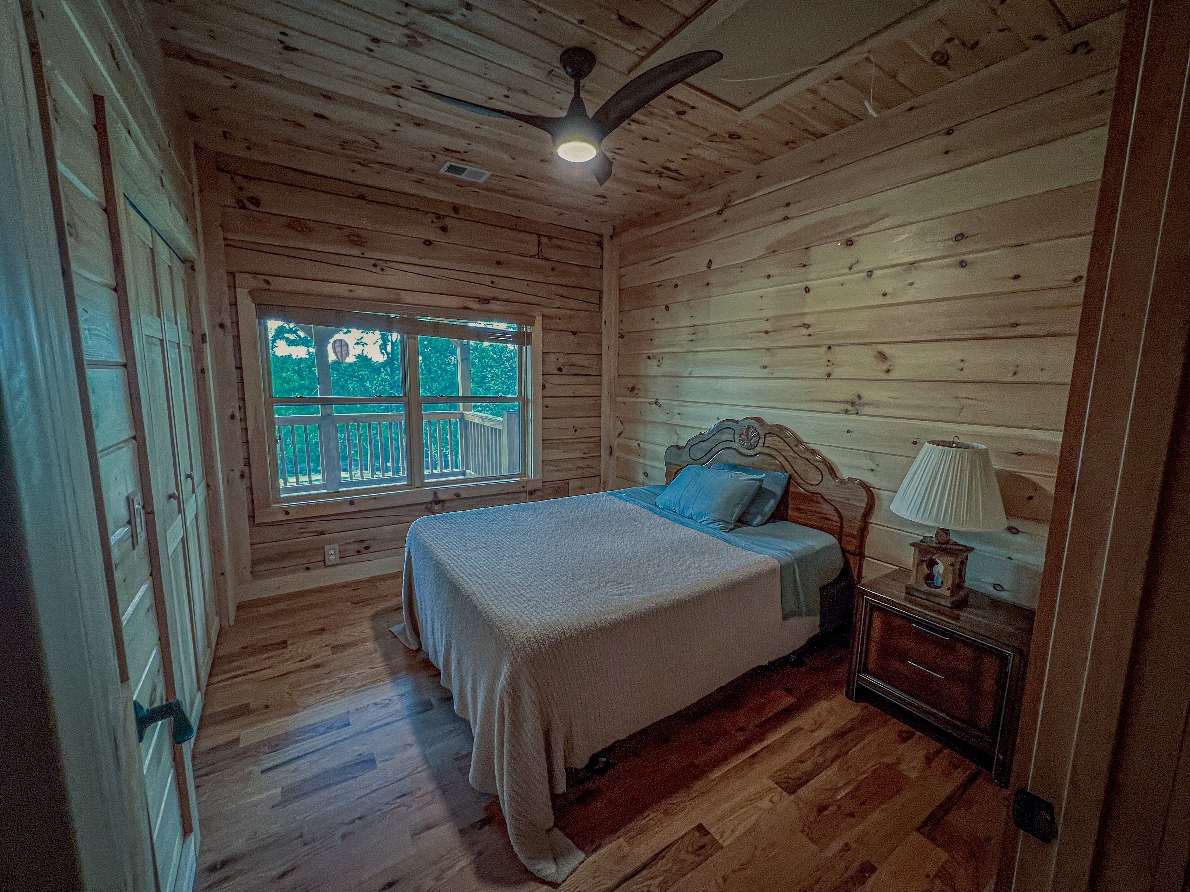 Quarto de cabana de madeira com uma cama, uma mesa de cabeceira, uma luminária e uma janela com vista para um deck.