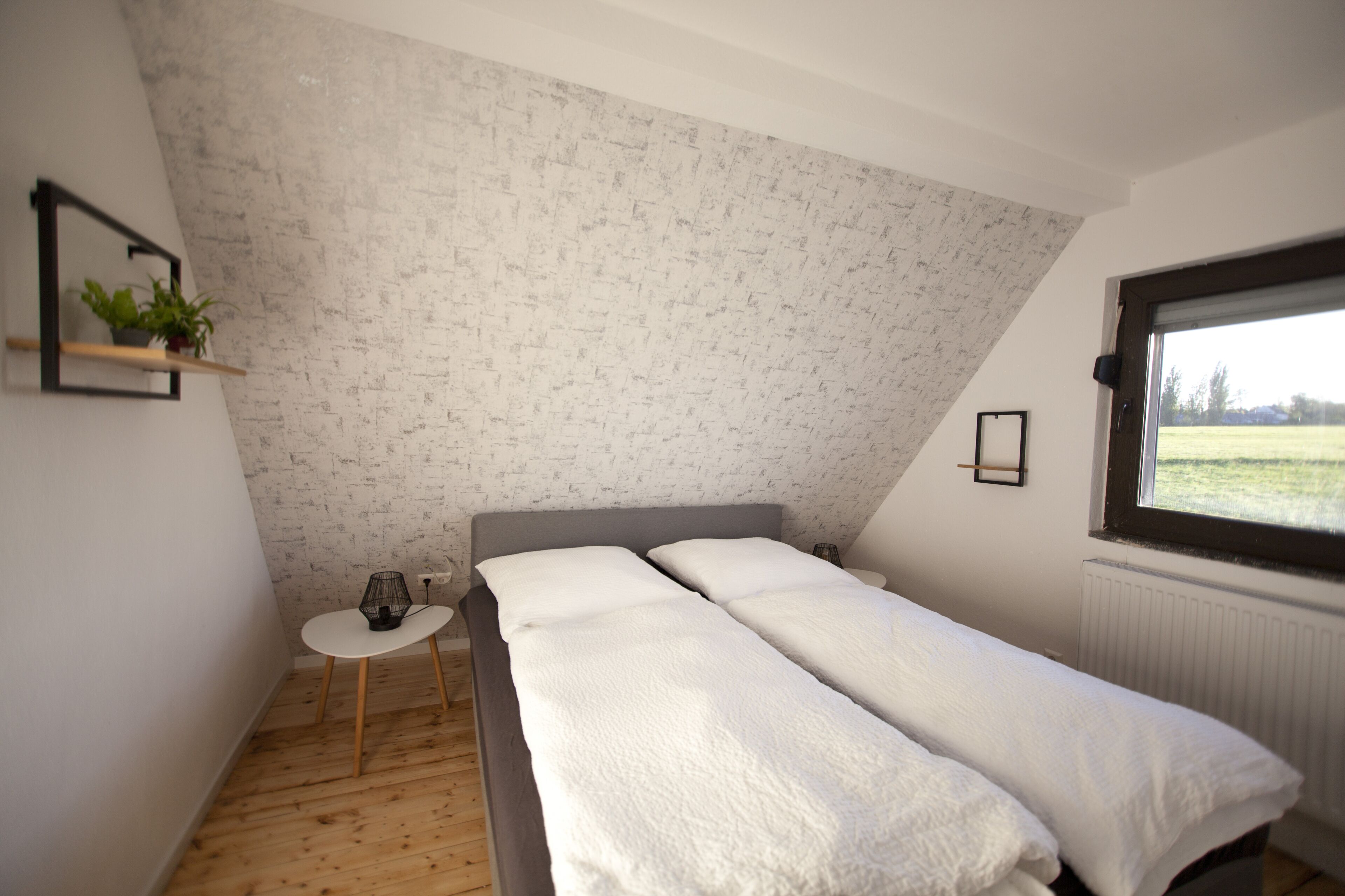 Een slaapkamer met schuine wand, een klein nachtkastje, een enkel bed met witte beddengoed, een raam met uitzicht op de omgeving, en een muur met een bakstenen patroon.