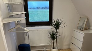 Um quarto com piso de madeira, um radiador, uma janela com vista para um campo, uma planta, um banquinho azul e um aparador branco.