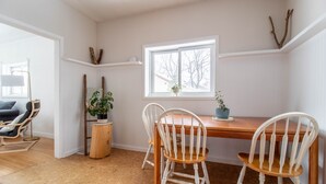A dining area with a wooden table and white chairs, a window with a view of trees, and a plant on the table.