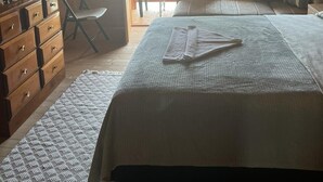 Une chambre confortable située sous les toits, avec un plafond en bois, un lit recouvert d’une couverture grise, une commode et une vue sur l’extérieur par une porte.