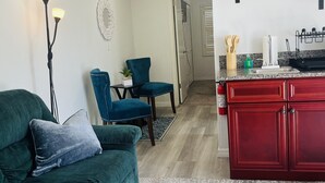 Ein Wohnzimmer mit einem grünen Sofa, einem blauen Sessel und einem kleinen Tisch mit einer Pflanze. Ein Küchenbereich mit roten Schränken und einer Arbeitsplatte ist ebenfalls zu sehen.